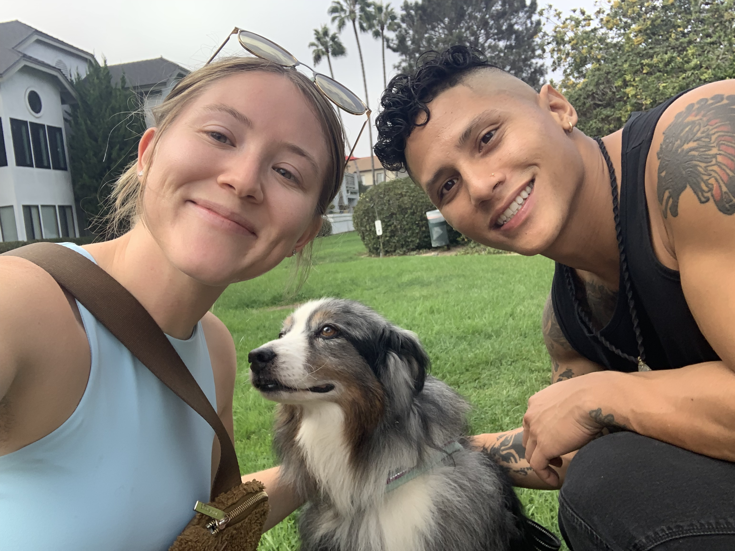 Two young adults, a woman and a man, smiling and taking a selfie with a dog on a grassy area in front of residential buildings.