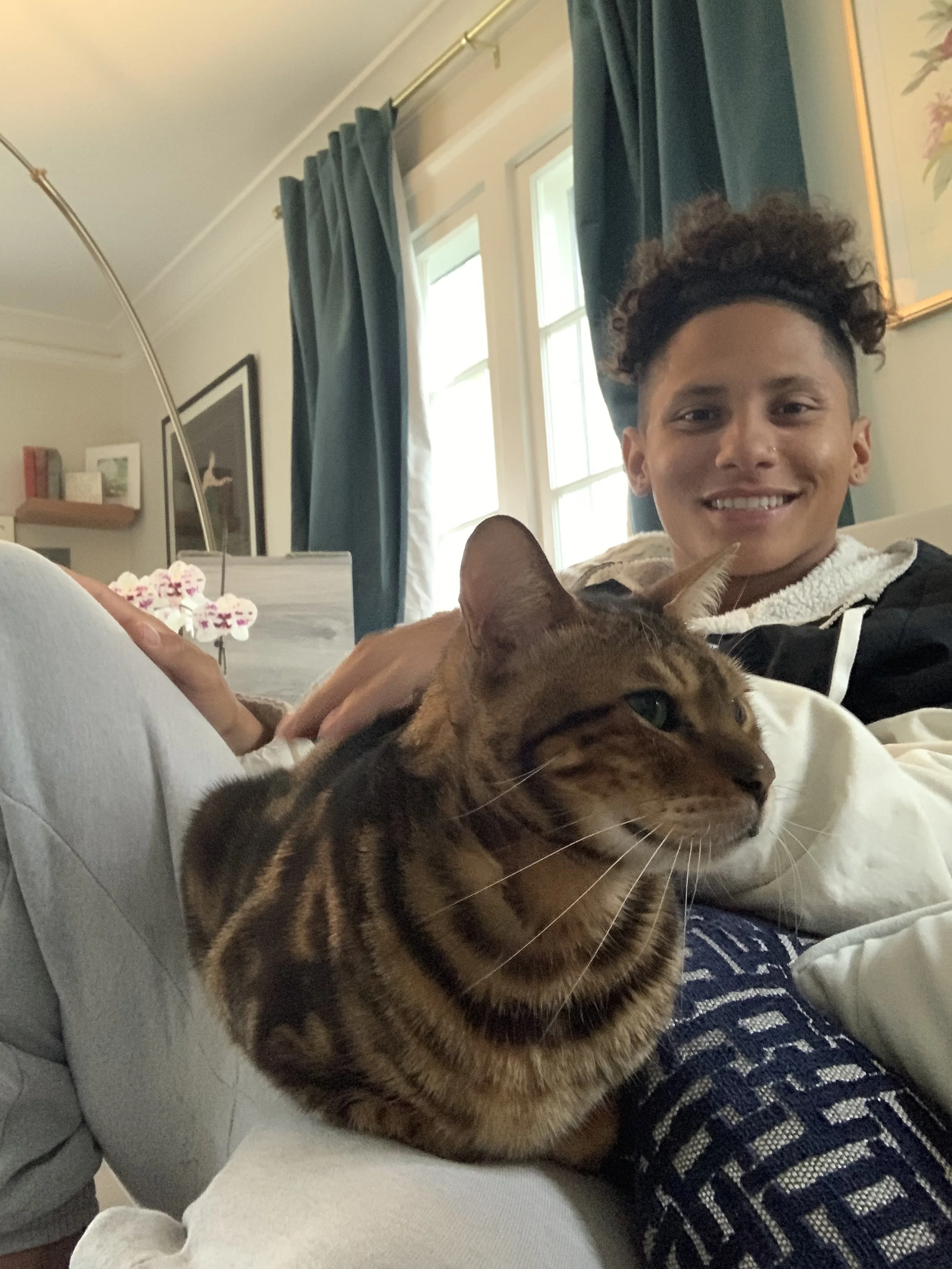 A person lying on a couch with a Bengal cat resting on their lap, smiling at the camera in a cozy living room with blue curtains and framed artwork on the wall.