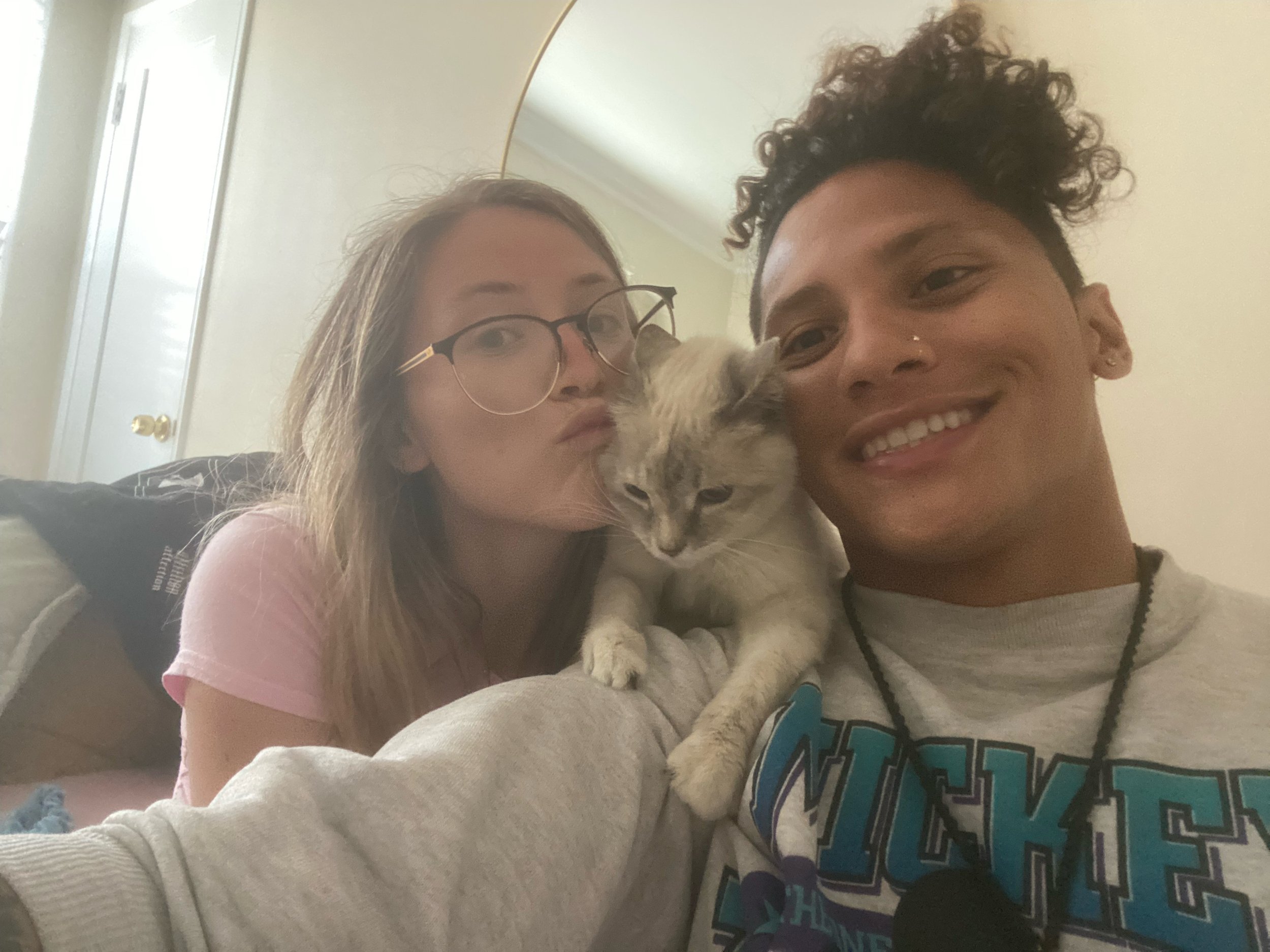 Two women and a gray tabby cat taking a selfie indoors. The woman on the left with glasses is making a duck face, and the woman on the right with curly hair has a big smile. The gray cat is being held close to their faces, looking slightly downward.