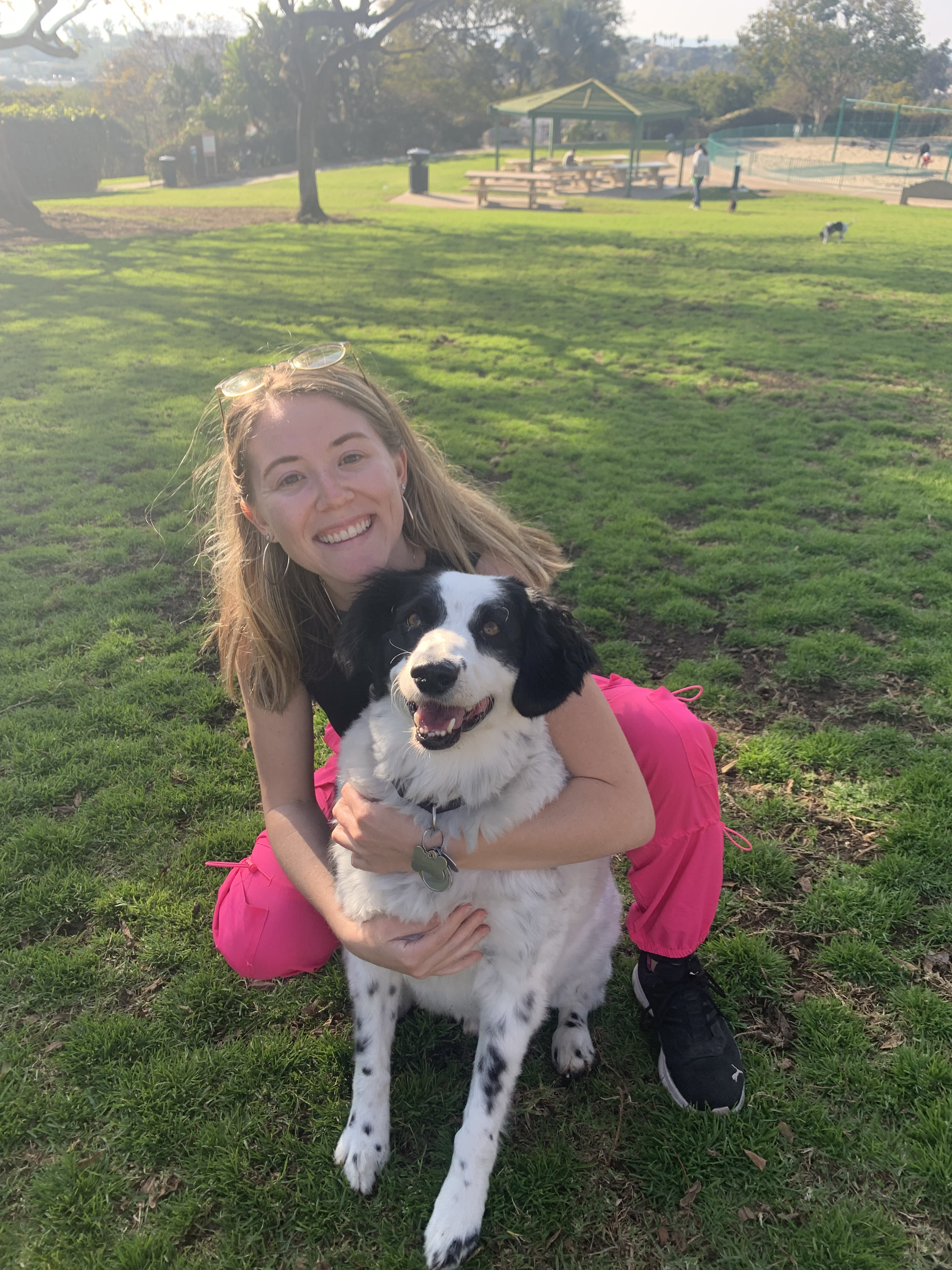 A woman with blonde hair, wearing a black shirt and pink pants, is kneeling on the grass and hugging a black and white dog. The park setting includes a picnic area with tables, a shelter, and a tennis court in the background, with two people and a small dog visible.
