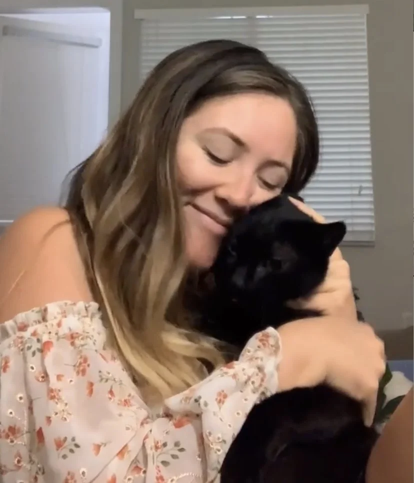 A woman with long brown hair and a floral off-shoulder top hugging a black cat, both appearing happy and content indoors.