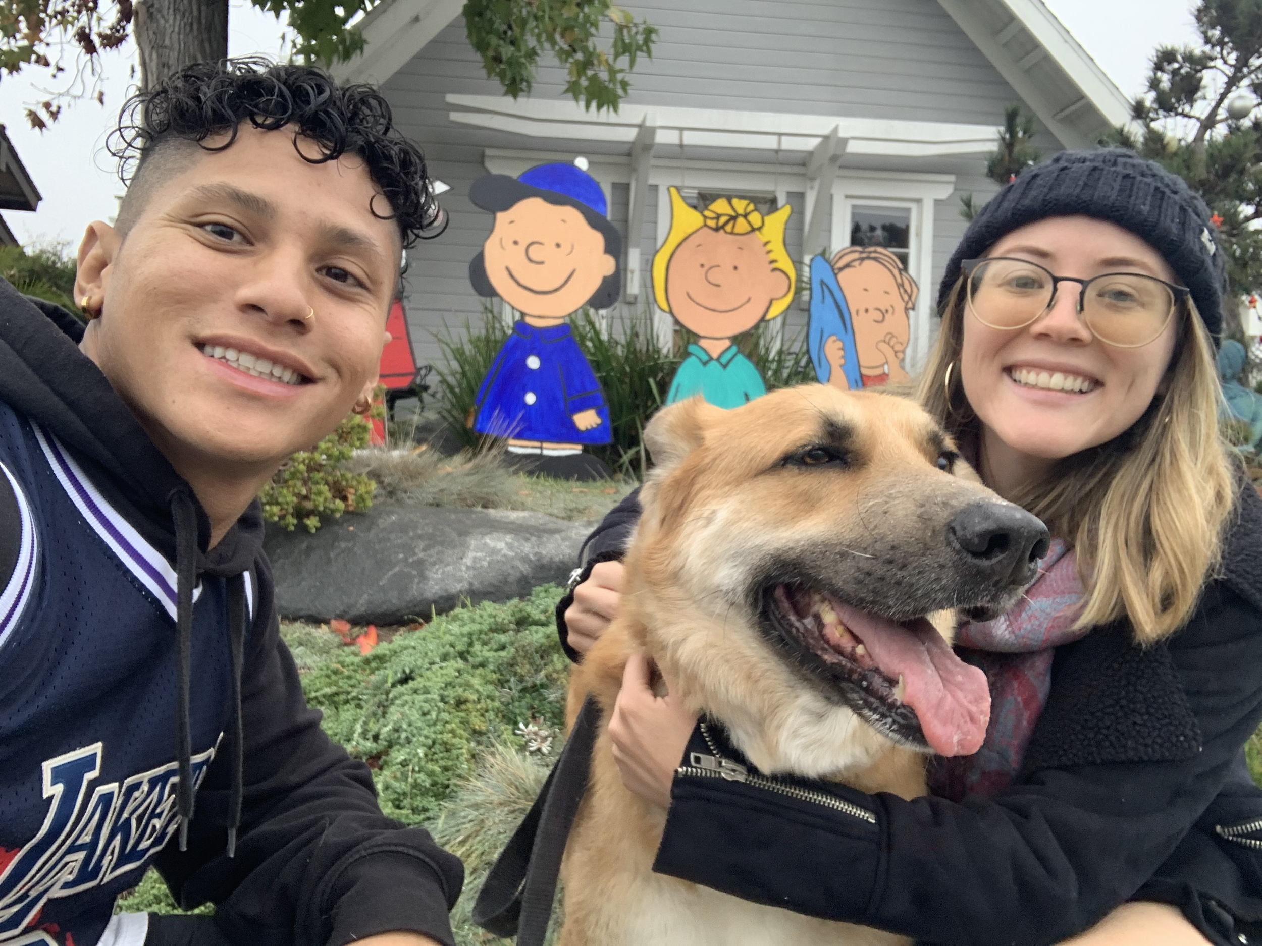 Two smiling people, one young man and one young woman, are taking a selfie with a happy dog. The background features cutout characters from the Peanuts comic strip, including Lucy, Sally, and Charlie Brown, in front of a house with plants and rock la