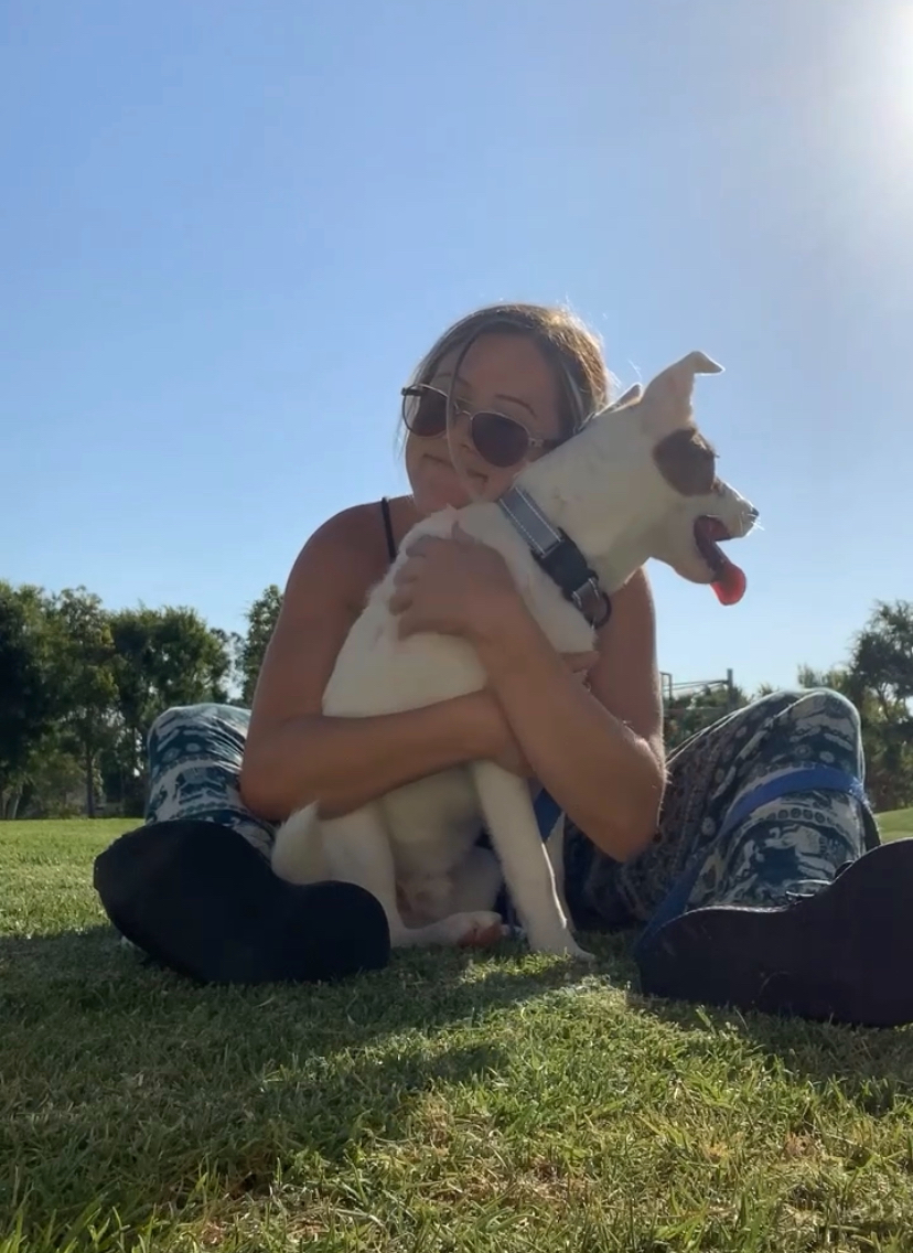 A woman with sunglasses sitting on the grass, hugging a white dog with brown spots, outdoors on a sunny day.