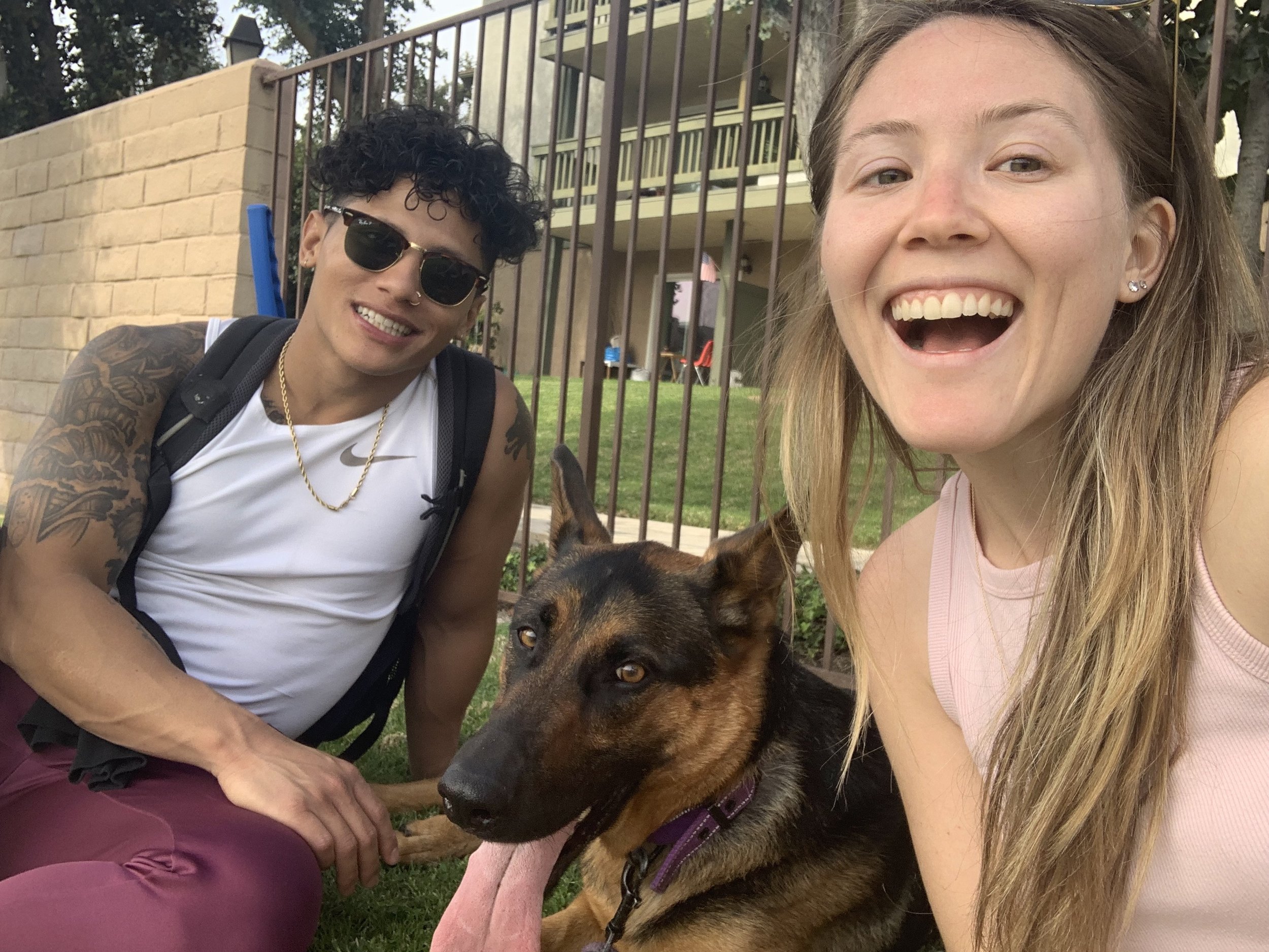 Two smiling women and a German Shepherd dog sitting on grass in front of a fence. One woman has curly dark hair, sunglasses, and tattoos on her arms, wearing a white tank top and maroon pants. The other woman has long blonde hair and is wearing a pin