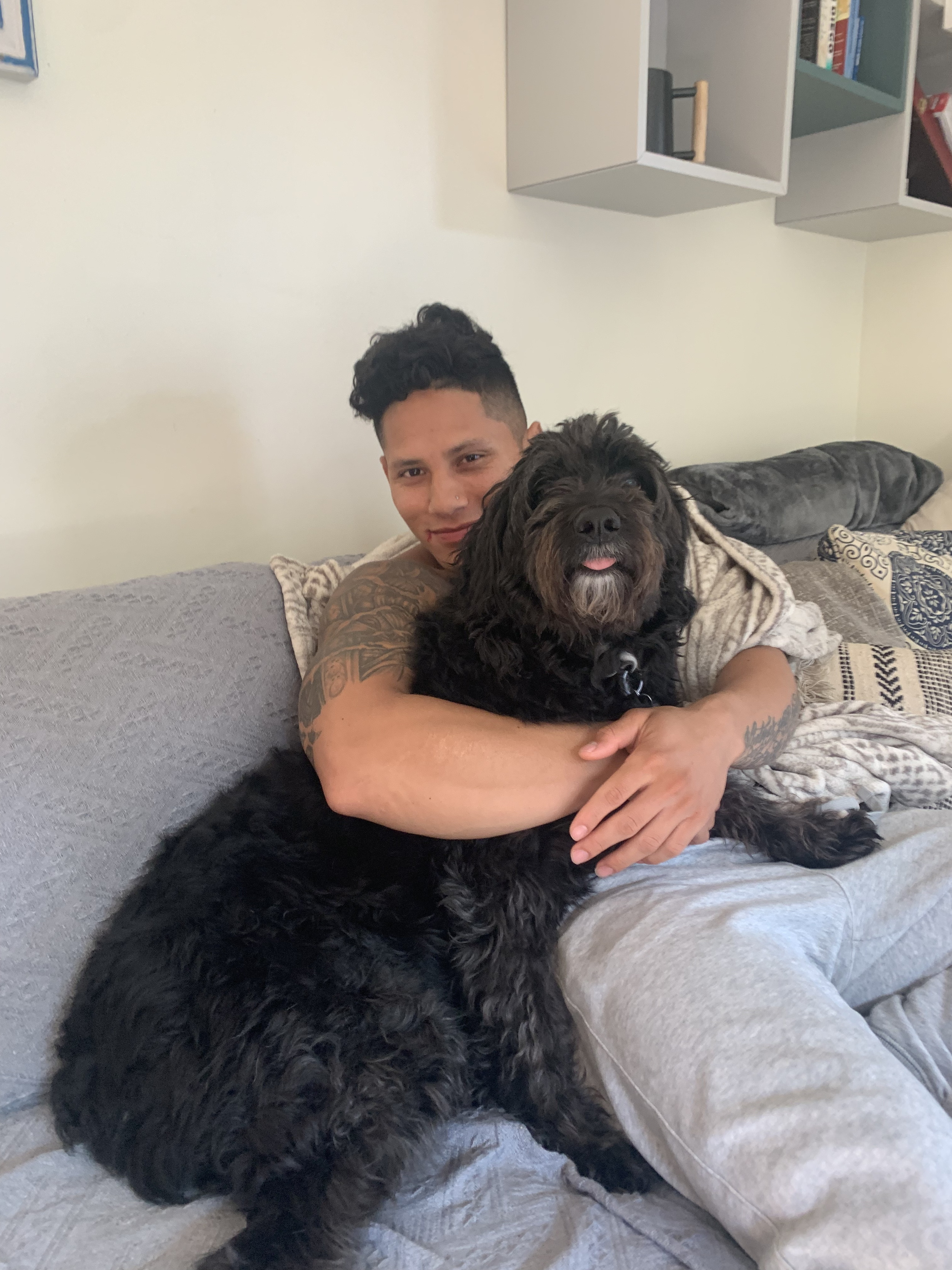 A person with short curly hair and tattoos hugging a black fluffy dog with its tongue out on a gray couch in a living room.