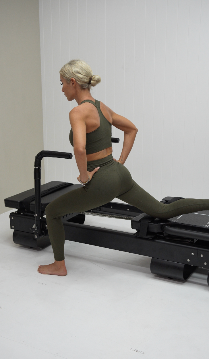 Woman with blonde hair in a bun wearing olive green workout leggings and sports bra, stretching on a Pilates reformer machine in a room with white walls.