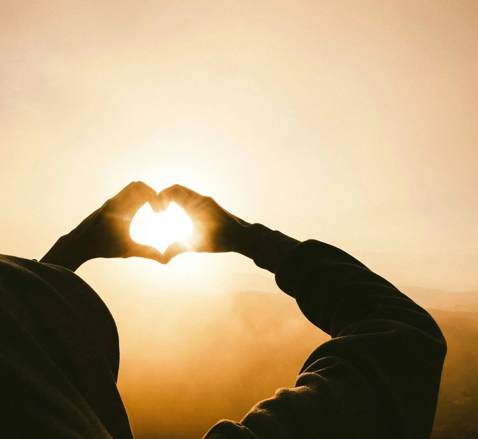 Person forming a heart shape with hands around the sun during sunset or sunrise.