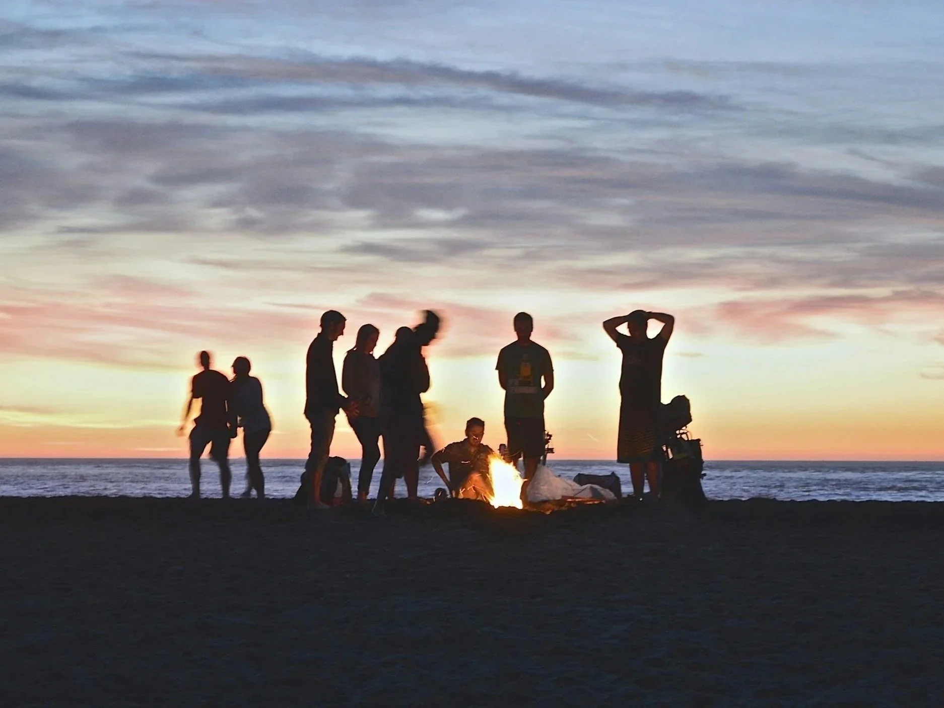 Group of people gathered around a campfire on a beach during sunset, with some standing and others sitting.