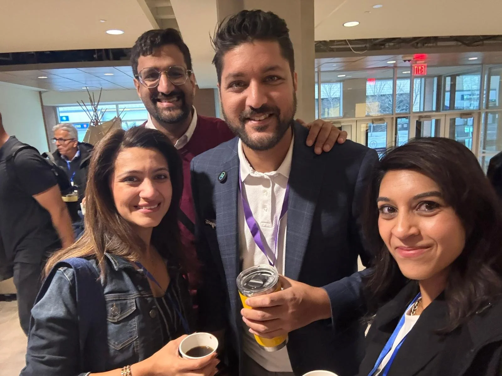 Four people smiling at an indoor event, holding coffee cups, with a man in a suit at the center. Two women are on either side, and a man is behind them, all looking at the camera.