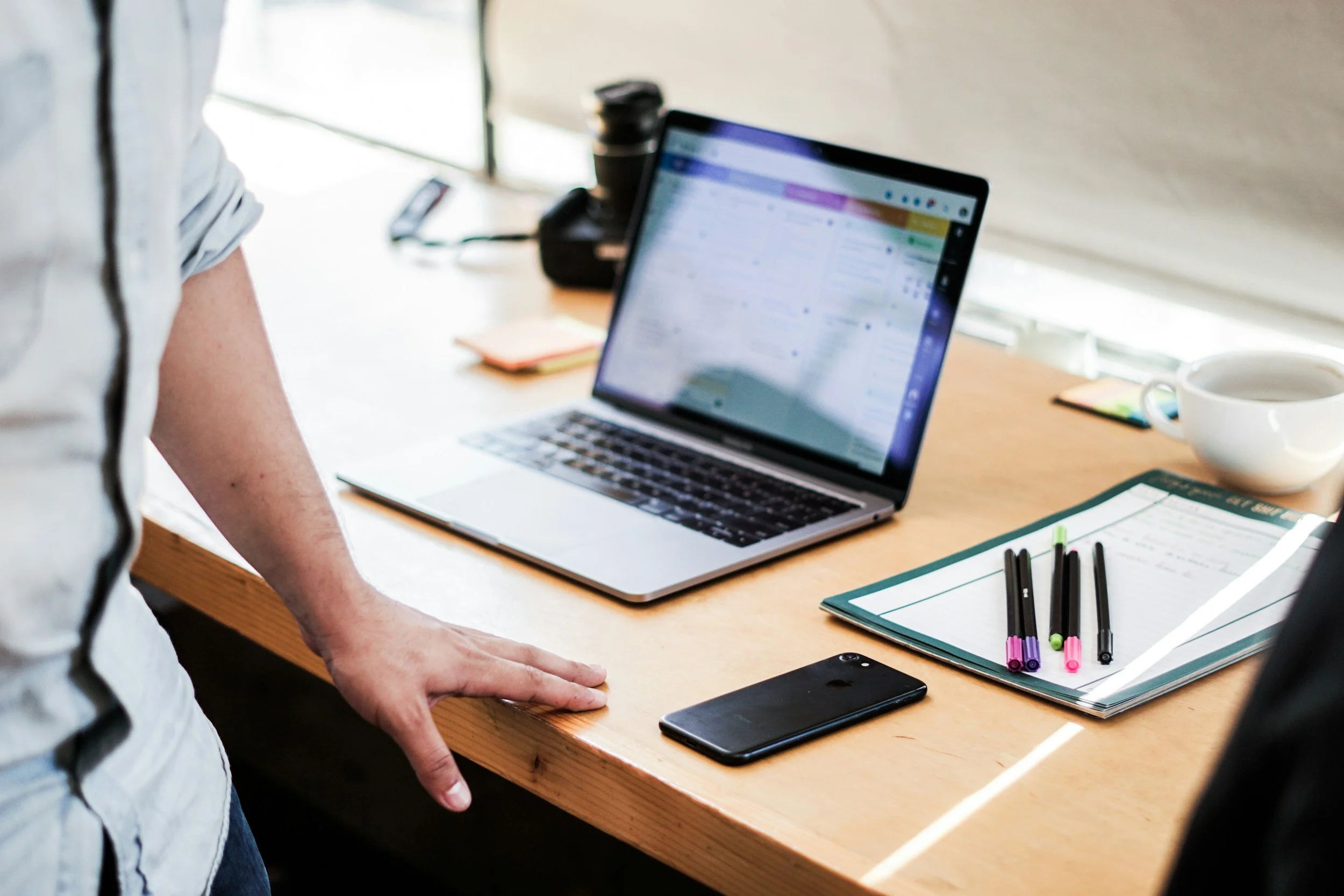 A person standing next to a desk with a MacBook laptop, a smartphone, a coffee mug, pens on notebooks, and a camera on the desk.