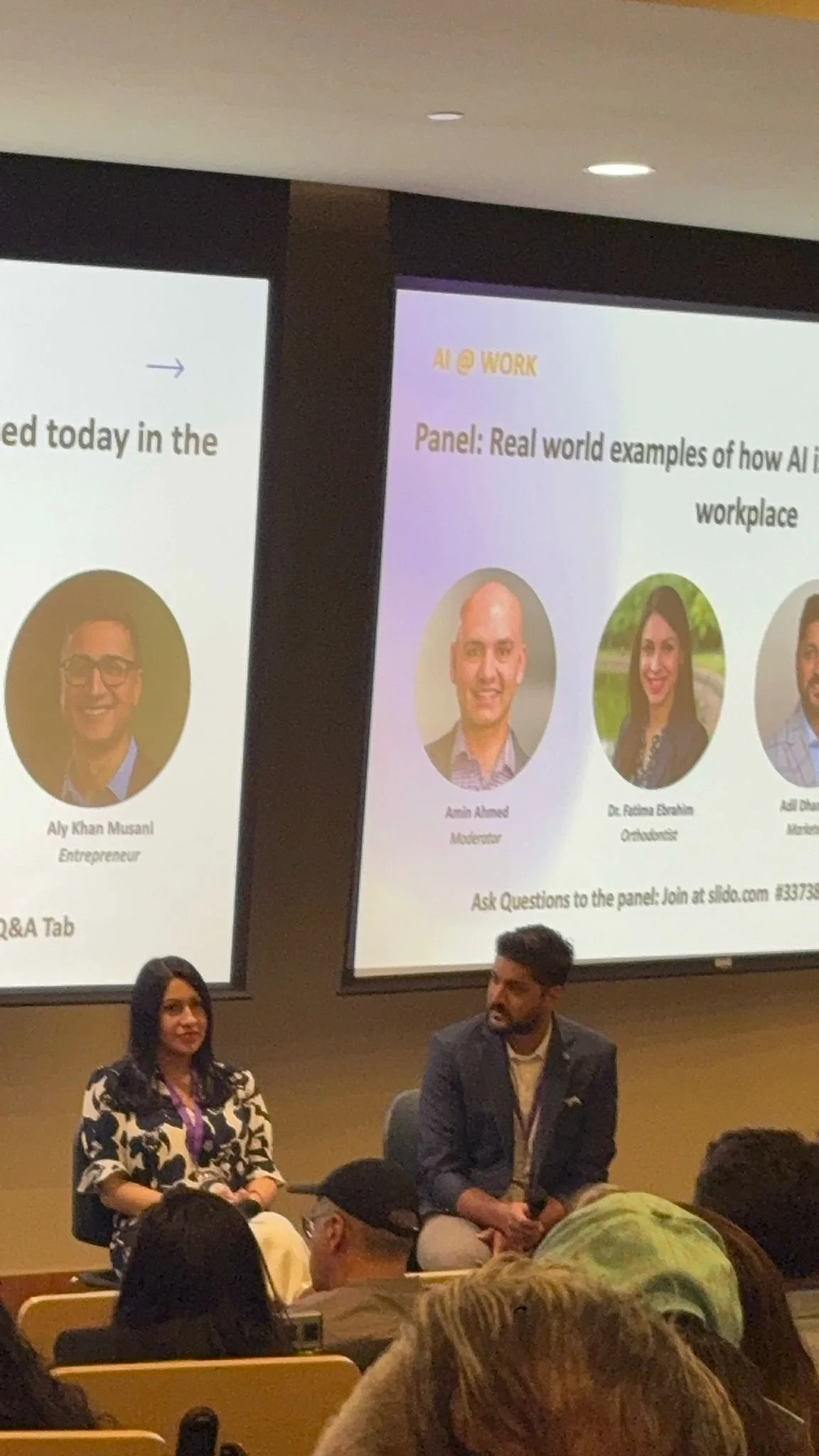 A panel discussion at a conference with two people seated in front of large screens displaying information about AI at work. The woman on the left has long black hair, is wearing a black and white patterned dress, and appears to be listening. The man