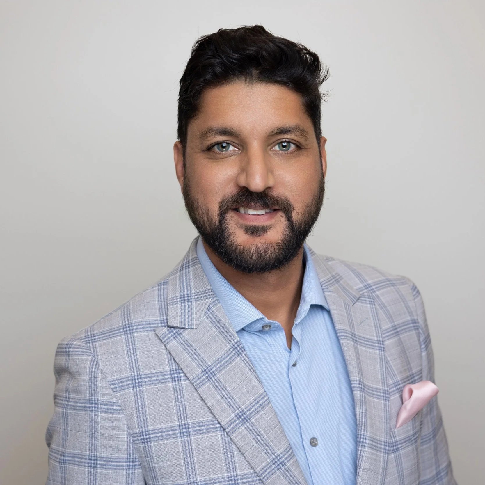 Headshot of a man with dark hair and beard, dressed in a light checkered blazer and blue shirt, smiling against a plain light background.