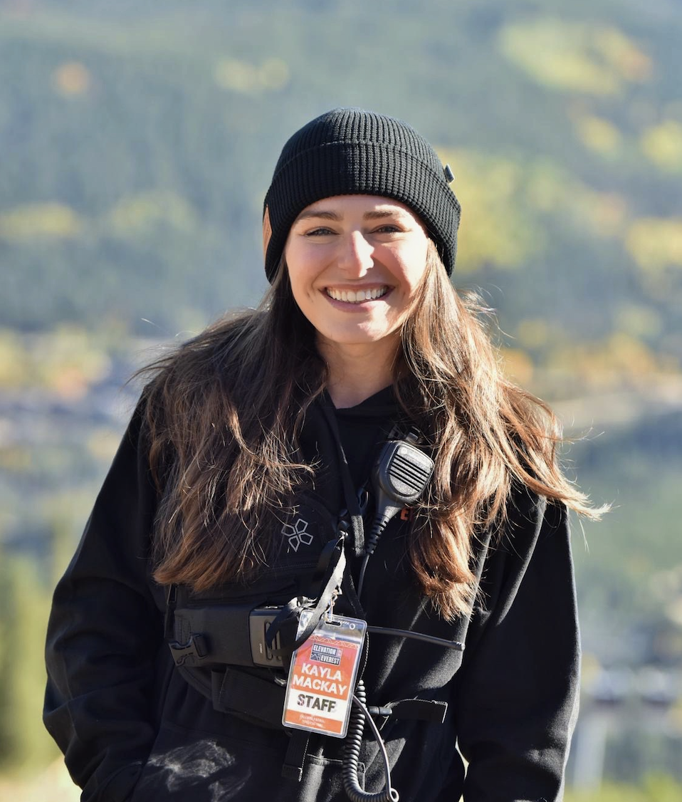 Smiling woman wearing a white cap with Everest logo, black jacket, and a radio microphone clipped to her collar, outdoors in a natural setting.