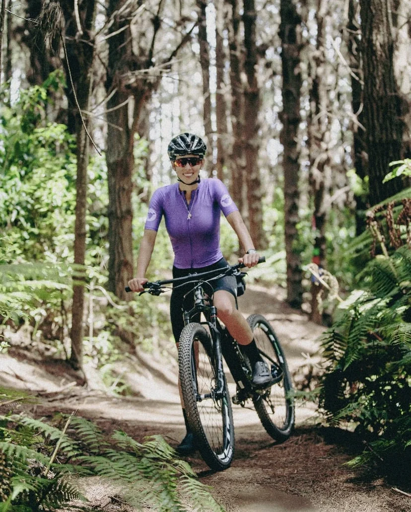A woman riding a mountain bike on a trail through a forest, wearing a purple cycling jersey, helmet, and sunglasses.