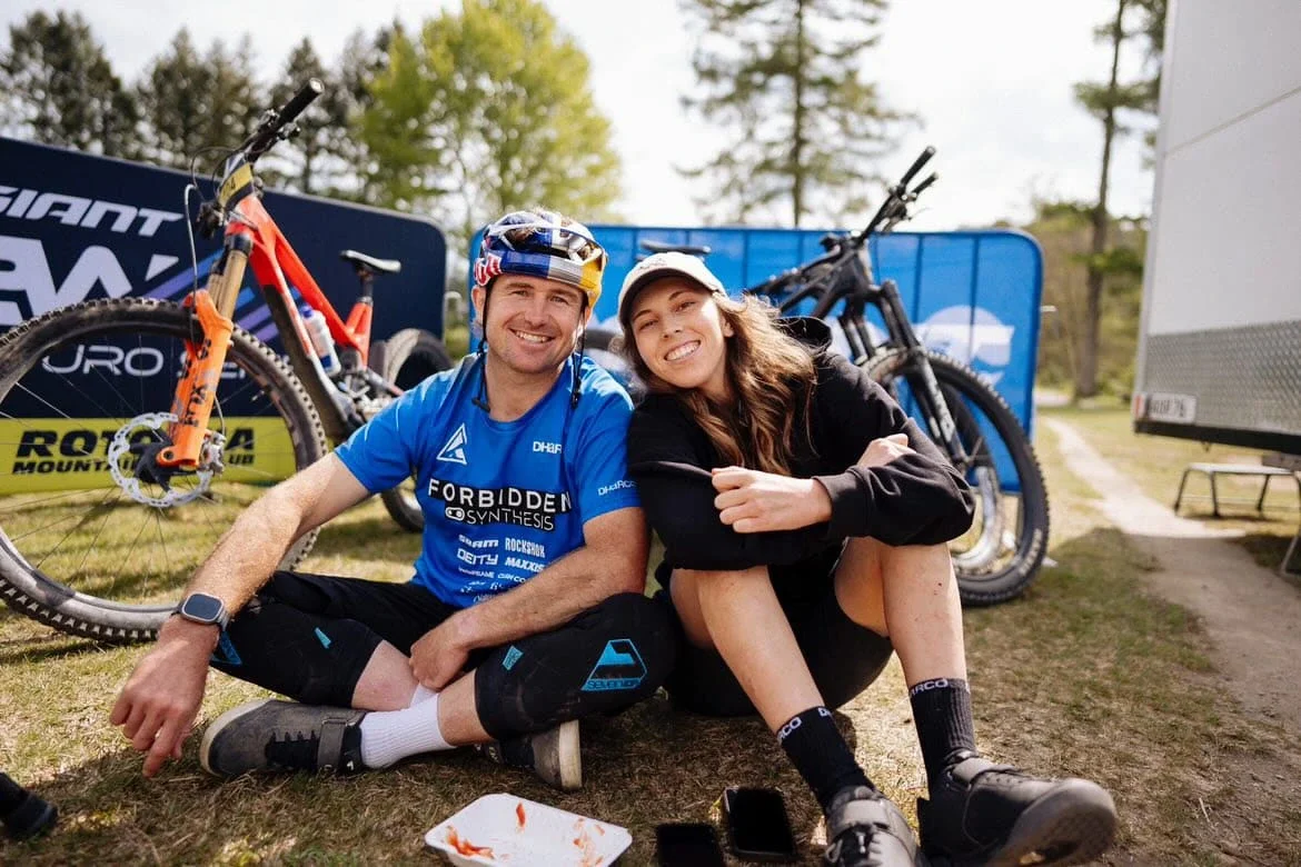 Two smiling mountain bikers sitting on the ground with their bikes behind them, surrounded by banners and trees.