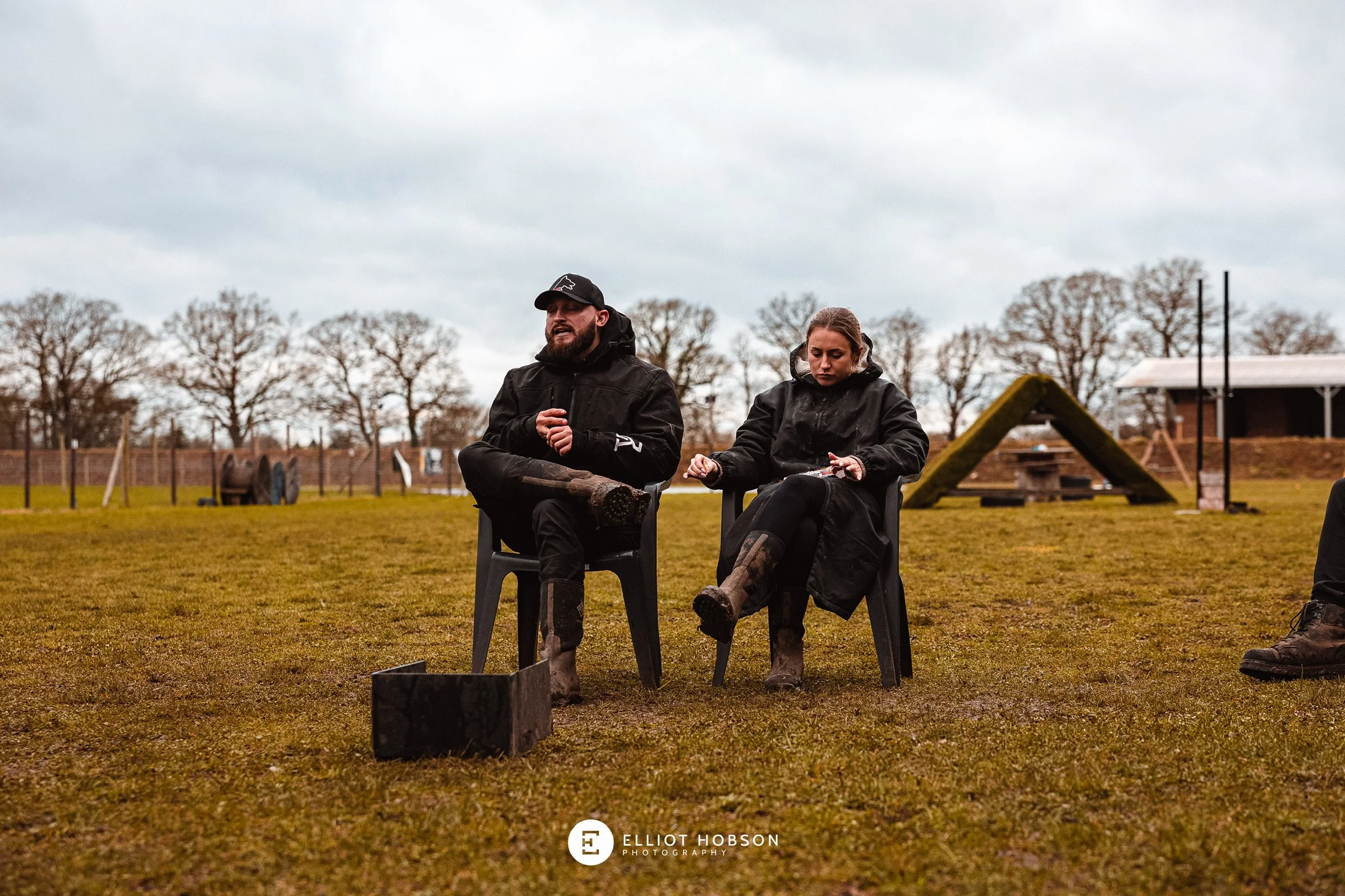 Two people sitting on black plastic chairs outdoors on a cloudy day, with trees in the background and worn grass, dressed in dark outdoor clothing and rubber boots, one man is talking, the woman is looking down at her lap.