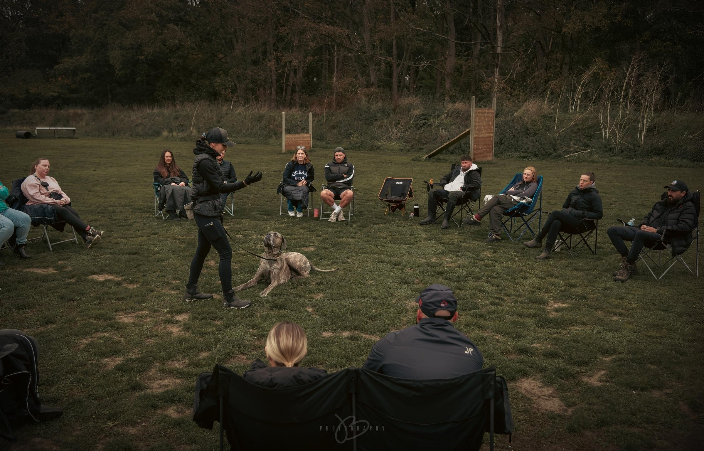 A group of people seated in chairs outdoors, watching a dog training demonstration led by a trainer with a guide dog on a leash.