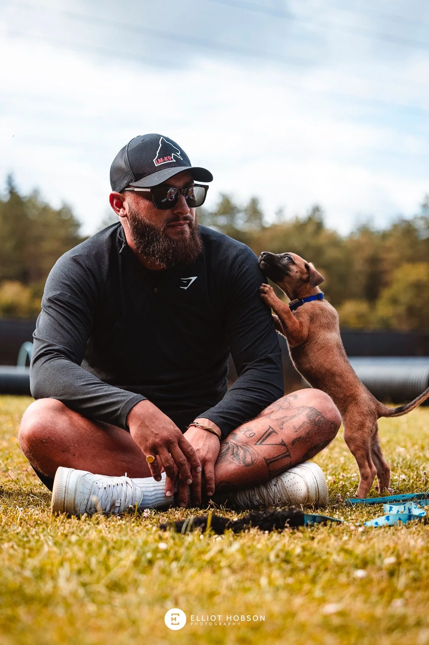 A man with a beard and tattoos sitting cross-legged on grass, wearing sunglasses, a black cap, and athletic clothing, playing with a small brown dog during daytime outdoor.