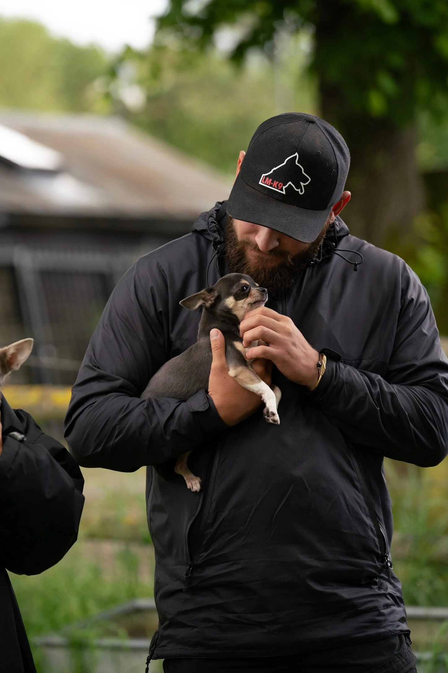 A man in a black jacket and black baseball cap holding a small chihuahua puppy outdoors, surrounded by green trees.