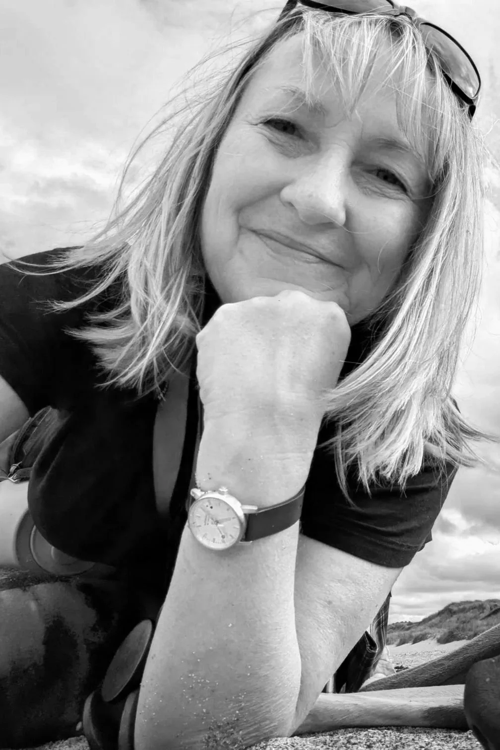 A woman with shoulder-length hair, smiling and resting her chin on her hand, wearing a watch and sunglasses on her head, outdoors against a cloudy sky.
