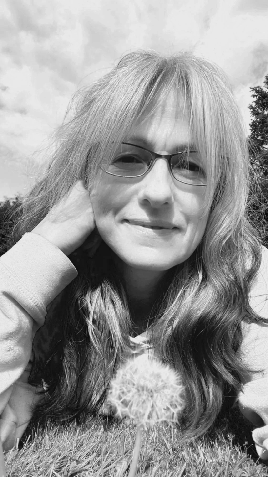 A woman with glasses laying on grass next to a dandelion, outdoor setting with clouds in the sky.