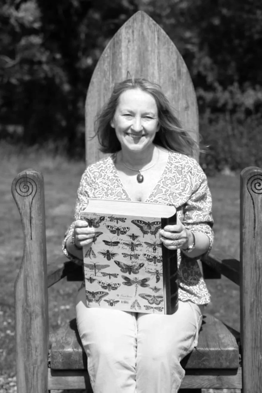 A smiling woman with shoulder-length hair, sitting outdoors on a large wooden chair with a tall, pointed backrest, holding a book decorated with various butterfly illustrations.