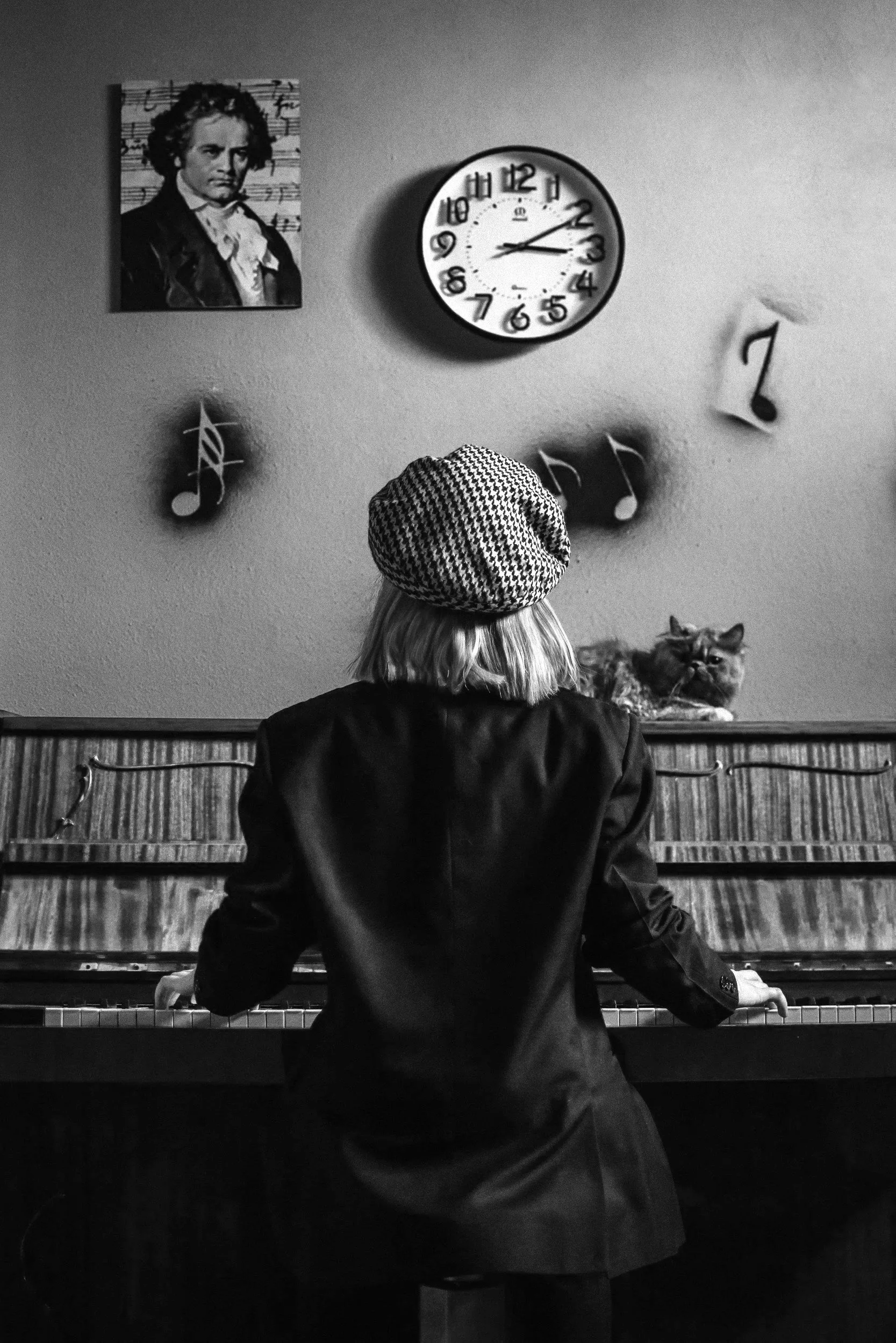 A person with shoulder-length hair wearing a houndstooth hat and a dark jacket, playing a piano with a cat sitting on top, in a room decorated with musical notes, a clock, and a portrait of Beethoven.
