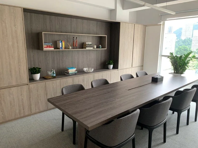 Partners Bay office workspace by Wan Fang Construction showing long wooden table, gray chairs, shelving unit with books and plant creating an organized atmosphere in Hong Kong Central.