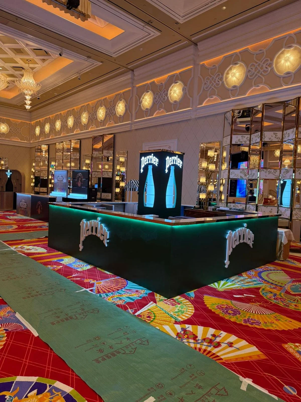 Perrier, an exhibition booth in Wanchai Hong Kong by Wang Fang Construction, features a sleek forest-green bar with an illuminated emerald top edge. A luminous display column glows with a light blue bottle silhouette creating futuristic brand environ