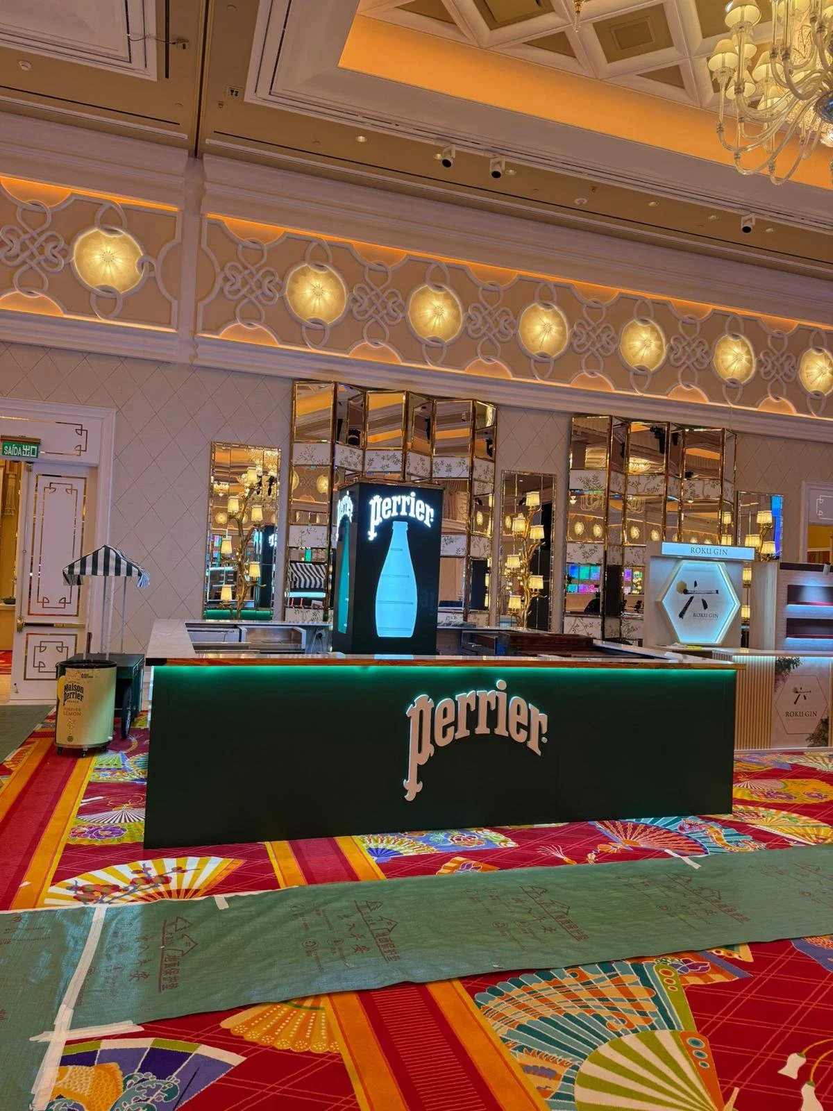 Perrier, an exhibition booth in Wanchai Hong Kong by Wang Fang Construction, features a sleek forest-green bar with an illuminated emerald top edge. A luminous display column glows with a light blue bottle silhouette creating futuristic brand environ