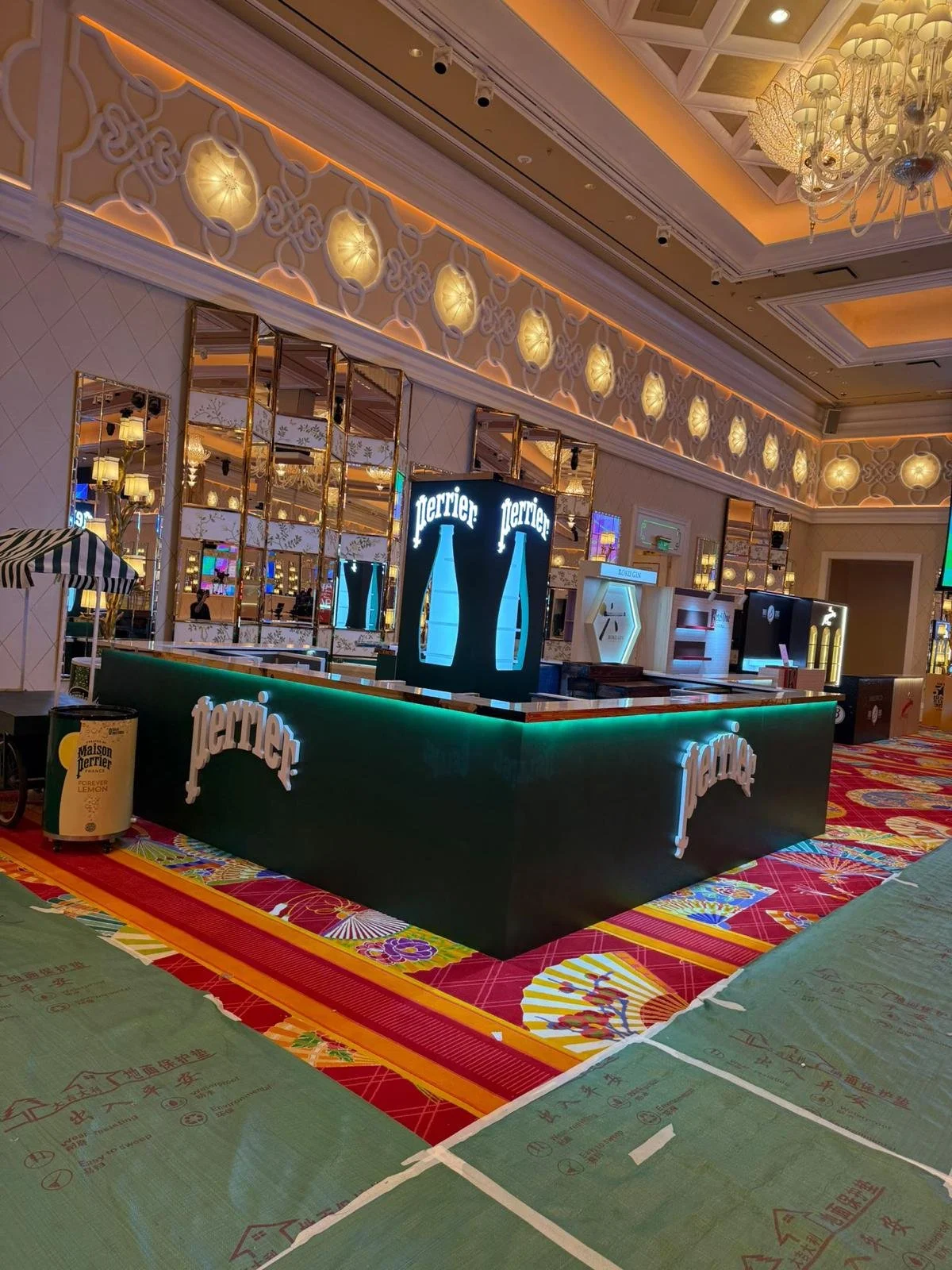 Perrier, an exhibition booth in Wanchai Hong Kong by Wang Fang Construction, features a sleek forest-green bar with an illuminated emerald top edge. A luminous display column glows with a light blue bottle silhouette creating futuristic brand environ