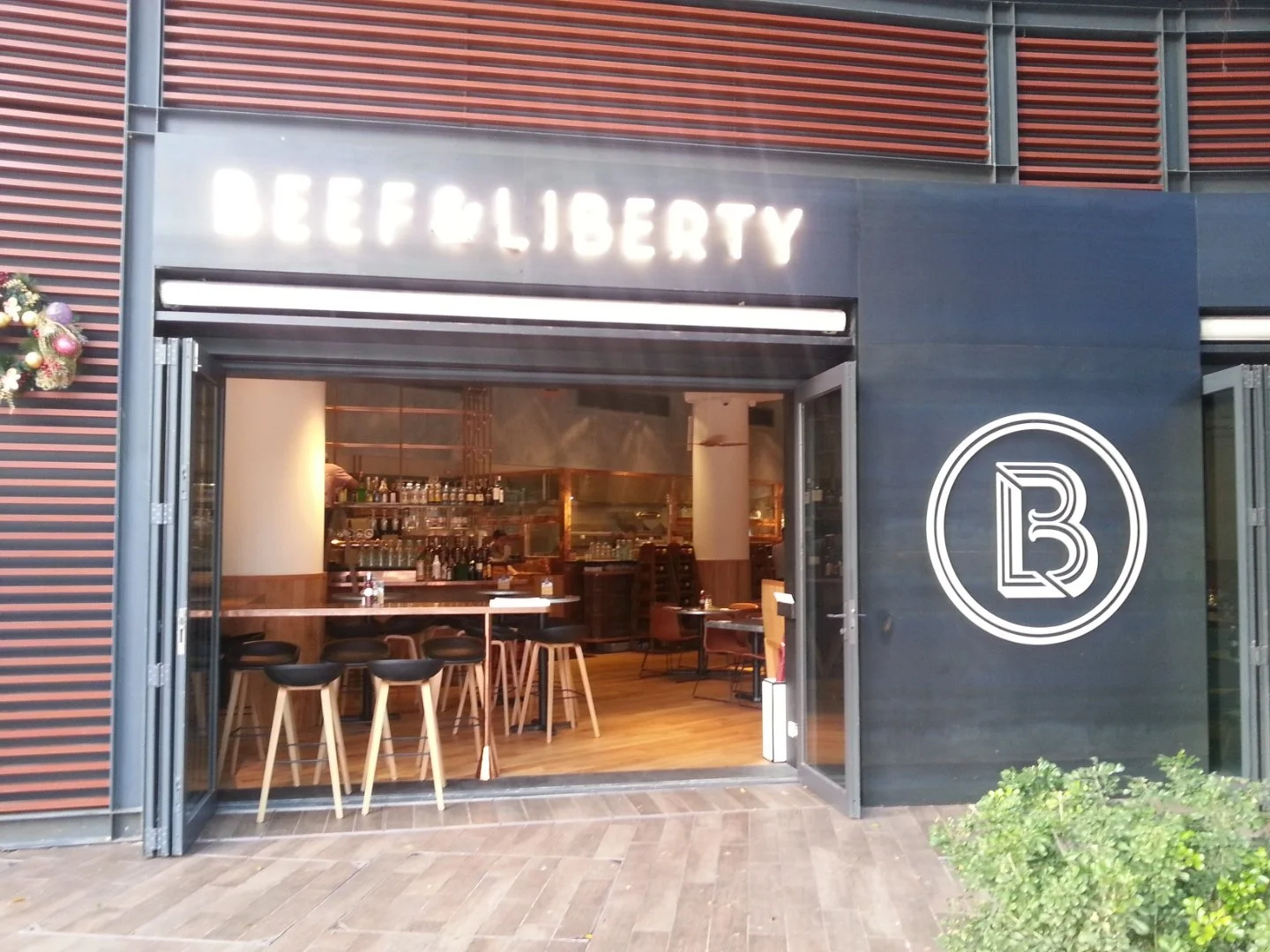 Beef & Liberty, a gourmet burger restaurant in Stanley Hong Kong by Wang Fang Construction, features a clean, modern interior. Visible through the glass doorway, the space is arranged with simple tables and chairs on a wooden floor. Bottles are lined