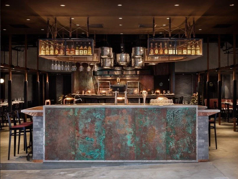 Rhoda modern restaurant with a central copper-toned bar counter by Wan Fang Construction, showing hanging pendant lights, shelving with bottles above, and a warm, dim lighting ambiance in Hong Kong, Sai Yin Pun.