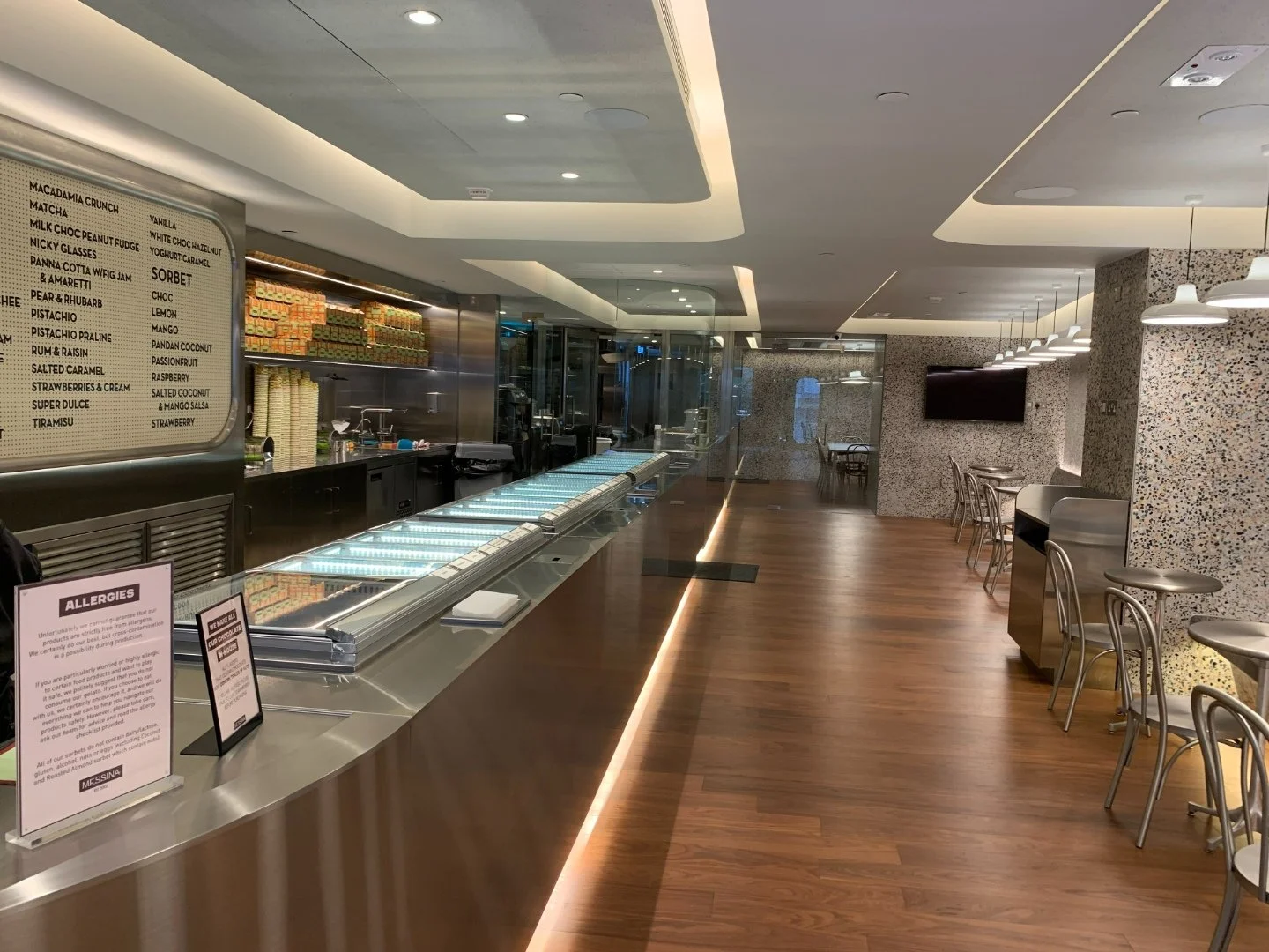 Messina Pattiserie interior by Wan Fang Construction features a warm wooden floor and a metallic counter displaying flavors. Minimalist metallic seating sits beneath pendant lighting. Decorative tiled walls and a large menu board complete the clean, 