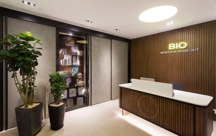 Bio Beauty Spa, a beauty boutique in Central Hong Kong by Wang Fang Construction, features a minimalist and professional interior. A stylish, vertically-paneled wooden reception desk with under-lighting is the centerpiece. Large light-colored tiles, 