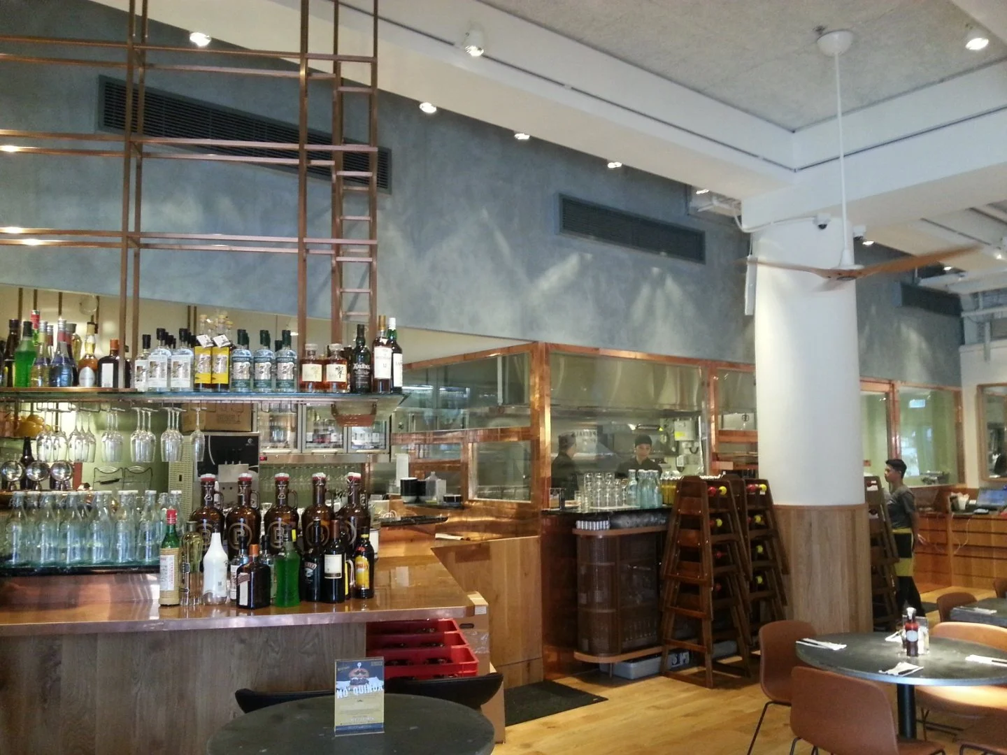 Beef & Liberty, a gourmet burger restaurant in Stanley Hong Kong by Wang Fang Construction, features an modern-chic interior. The foreground includes a bar counter with bottles and round tables, while a glass partition reveals a stainless steel kitch