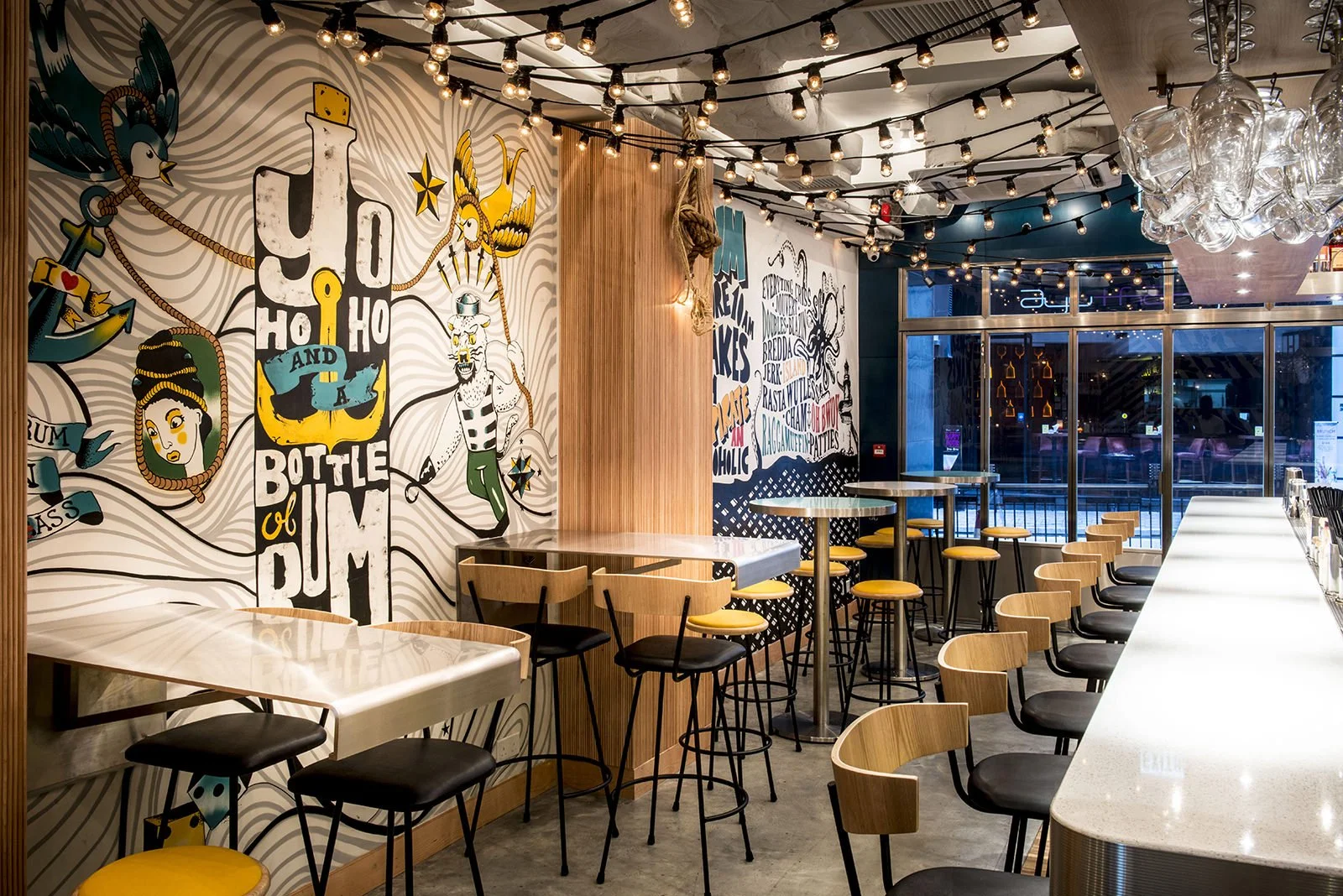 Rummin' Ting, a Caribbean-inspired bar and restaurant in Central Hong Kong by Wan Fang Construction, features a vibrant interior. A central, backlit bar displays bottles, surrounded by stools and round tables with distinctive yellow seats. The modern
