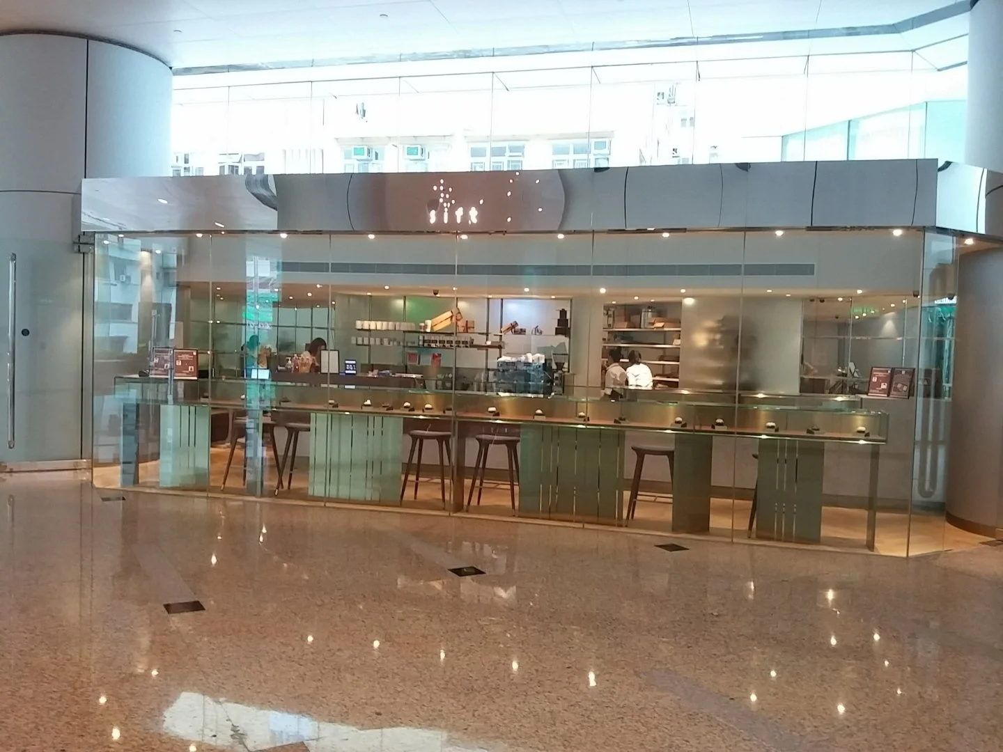 Sift, a modern patisserie in Quarry Bay Hong Kong by Wang Fang Construction, is a sleek glass kiosk within a bright, airy space. The long counter, with bar stools for seating, houses service appliances and display shelves. Neutral tones and integrate