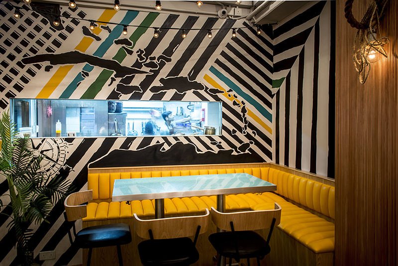 Rummin' Ting, a Caribbean-inspired bar and restaurant in Central Hong Kong by Wan Fang Construction, features a vibrant interior with a bold black and white striped wall. A bright yellow banquette provides cozy seating alongside a glass-topped table 