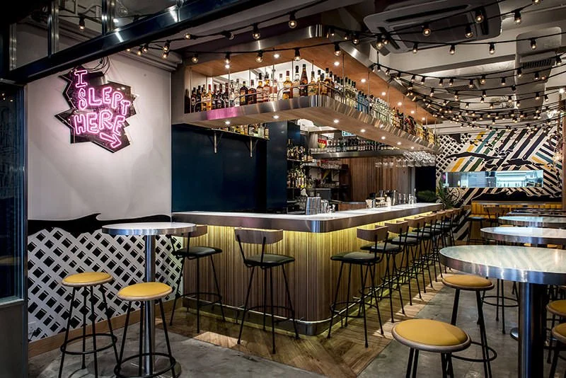 Rummin' Ting, a Caribbean-inspired bar and restaurant in Central Hong Kong by Wan Fang Construction, features a vibrant interior. A central, backlit bar displays bottles, surrounded by stools and round tables with distinctive yellow seats. The modern