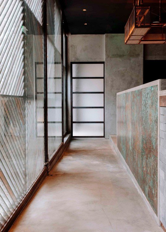 Rhoda modern restaurant by Wan Fang Construction, shows modern industrial corridor features a polished concrete floor and a dark ceiling. A slatted wall filters light from a large window, opposite a rustic copper-paneled wall in Hong Kong, Sai Yin Pu