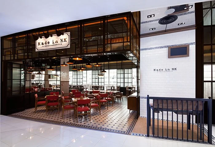 Made in HK, a modern restaurant in Hong Kong Kwun Tong by Wang Fang Construction features sign, black pendant lights, red leather chairs, and white-topped tables. A long wooden counter and shelves of colorful utensils adorn the back wall, while a pat