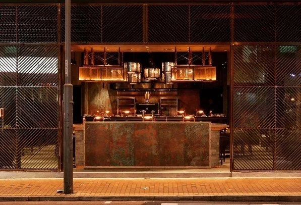 Rhoda modern restaurant with a central copper-toned bar counter by Wan Fang Construction, showing hanging pendant lights, shelving with bottles above, and a warm, dim lighting ambiance night view in Hong Kong, Sai Yin Pun.