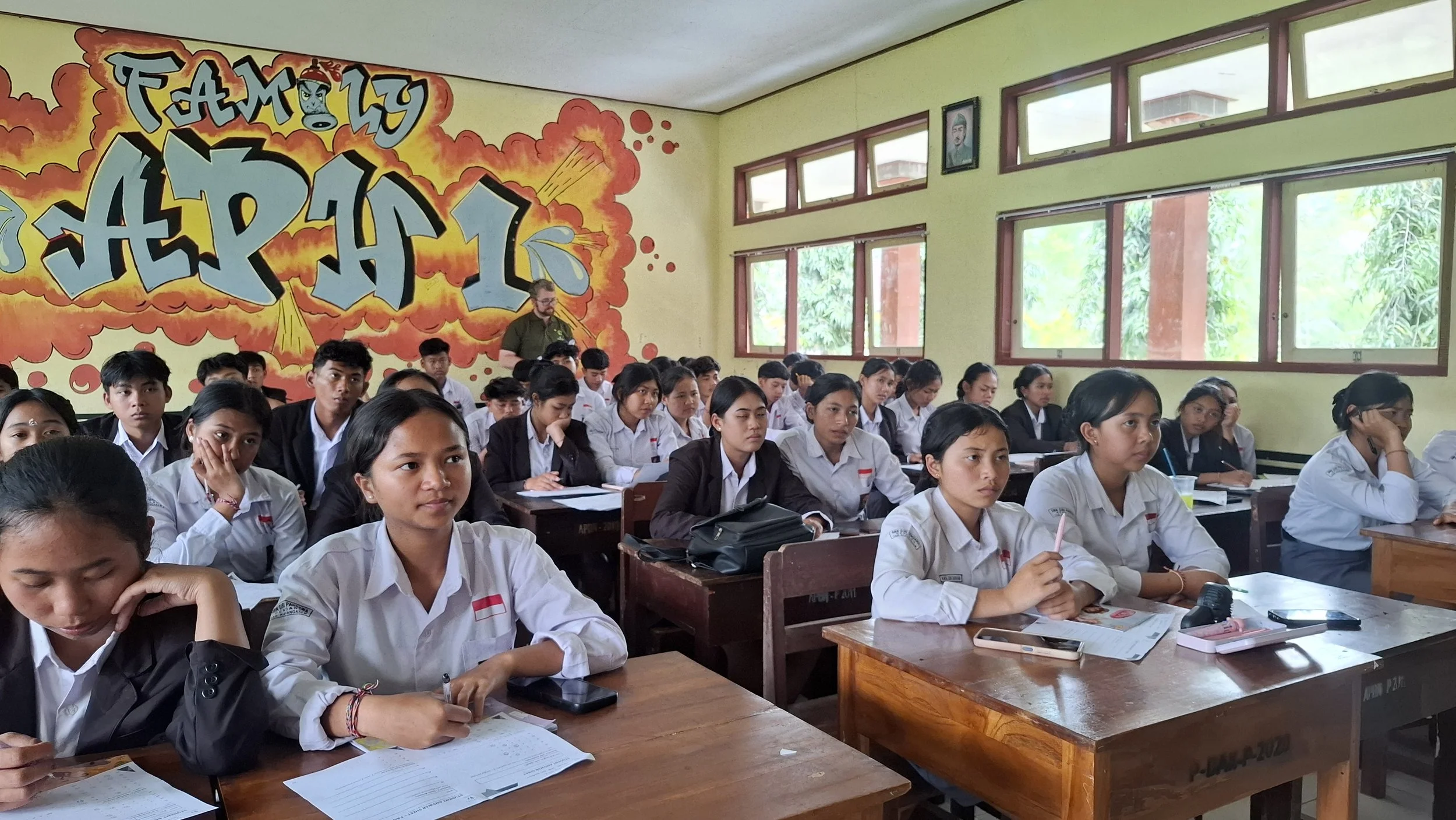 Classroom full of students dressed in white shirts and some in dark jackets, sitting at wooden desks, listening attentively, with a colorful graffiti-style wall that reads 'FAMILY' and other graffiti art, windows showing trees outside.