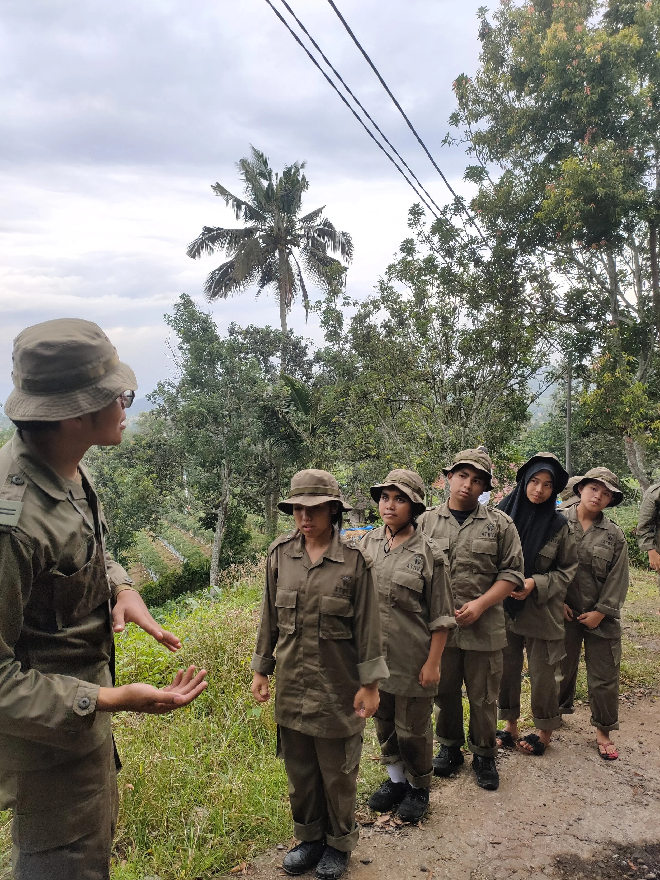 A group of young people in uniform standing outdoors on a dirt path, listening to an adult who is gesturing with his hands, with trees and sky in the background.