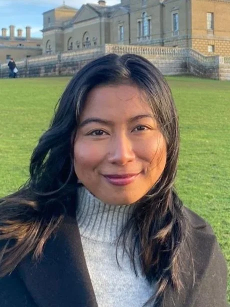 A young woman with shoulder-length dark hair smiling at the camera. She is indoors, with a background of wooden window frames and blurred outdoor scenery.