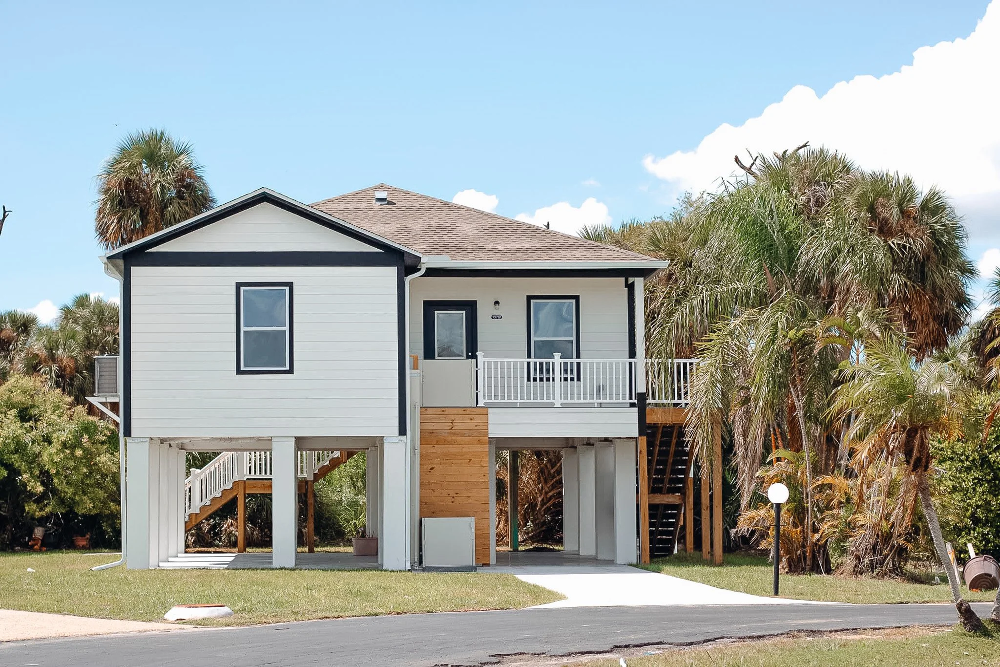 A raised beach house with white siding and black trim, a small front porch, and stairs leading to the entrance, surrounded by palm trees under a blue sky.