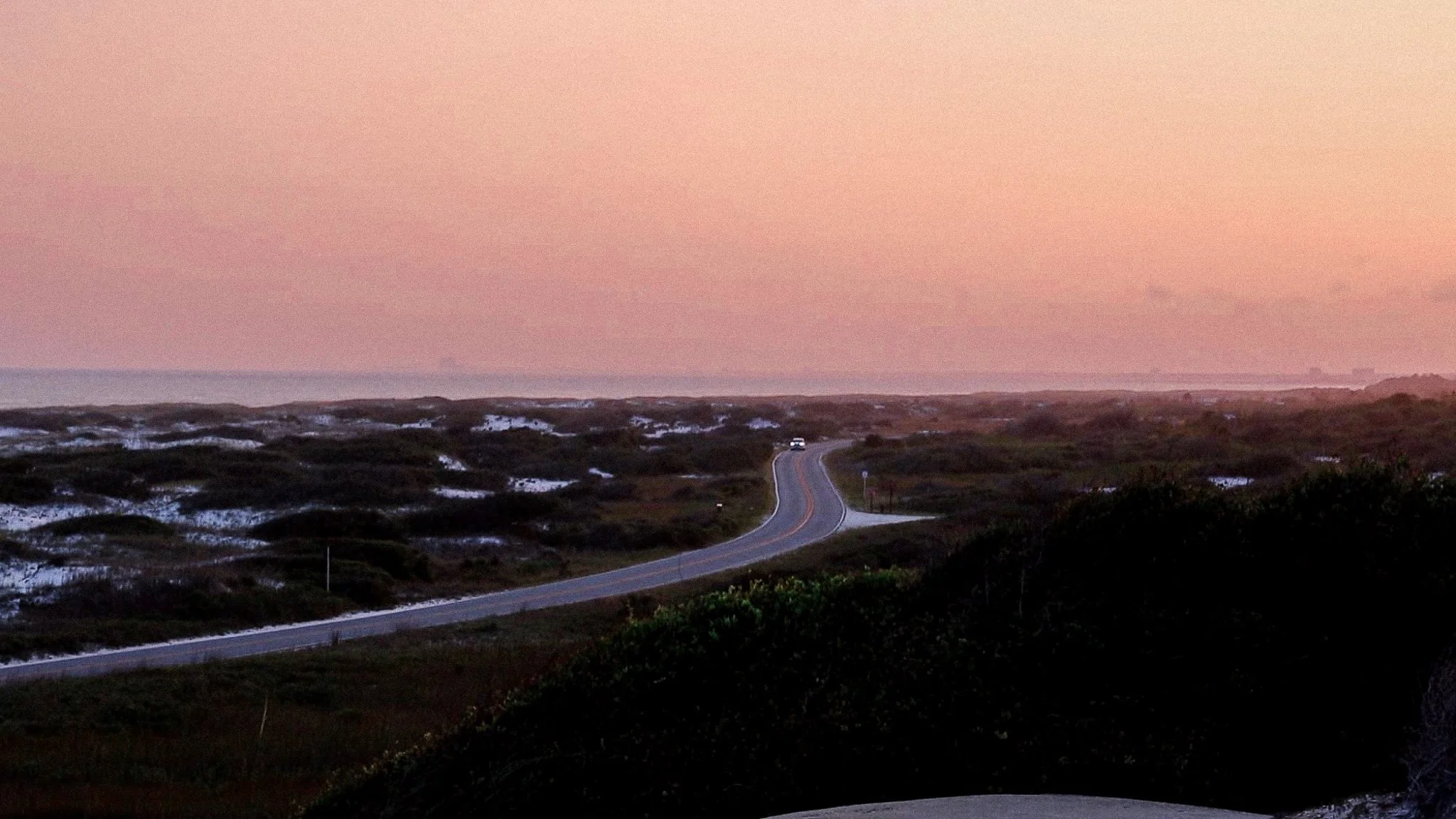 A winding road through coastal dunes at sunset with a car in the distance, pink sky and patches of snow on the dunes.
