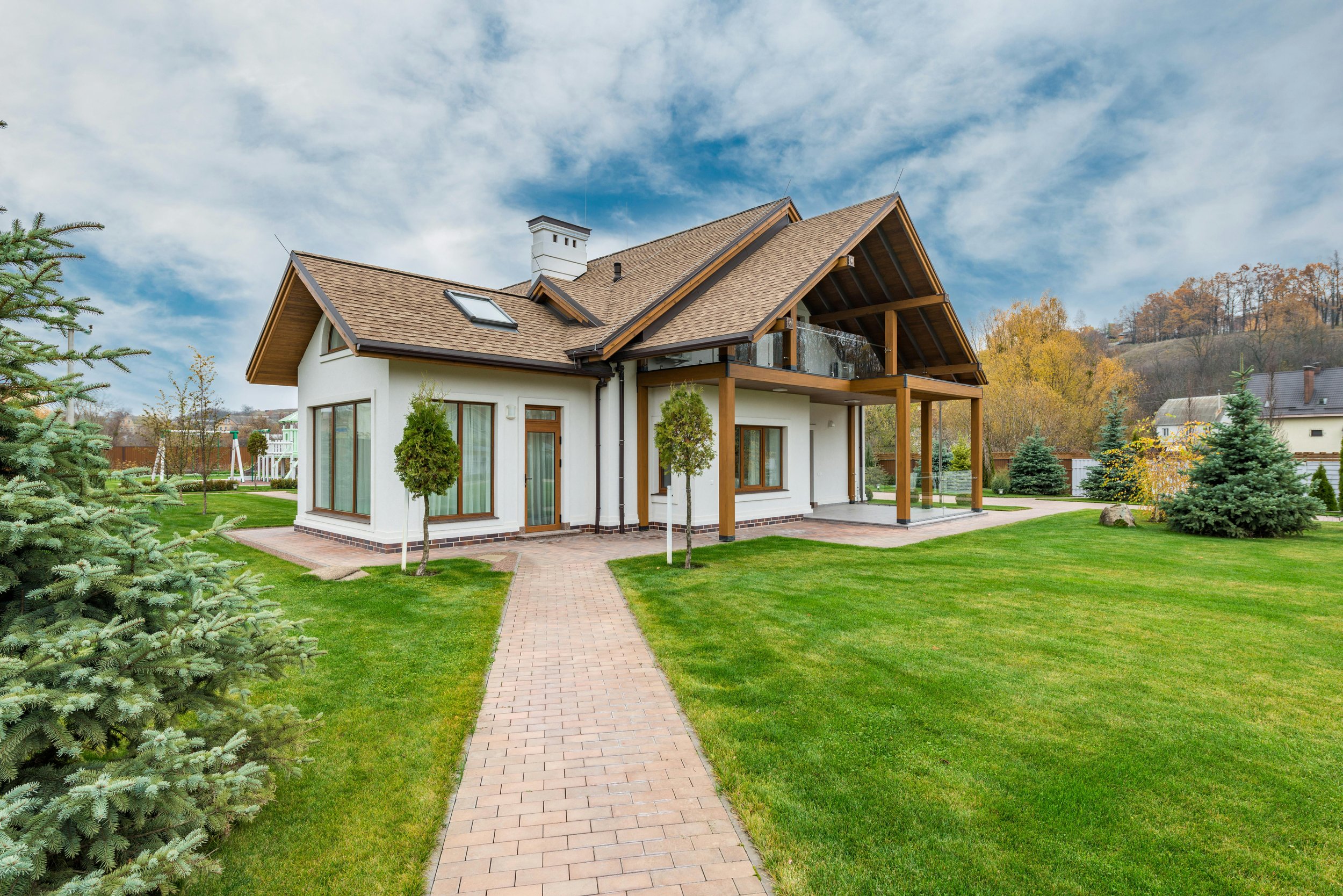 A modern house with a brown roof, white walls, and large windows, surrounded by a green lawn and trees, under a partly cloudy sky.