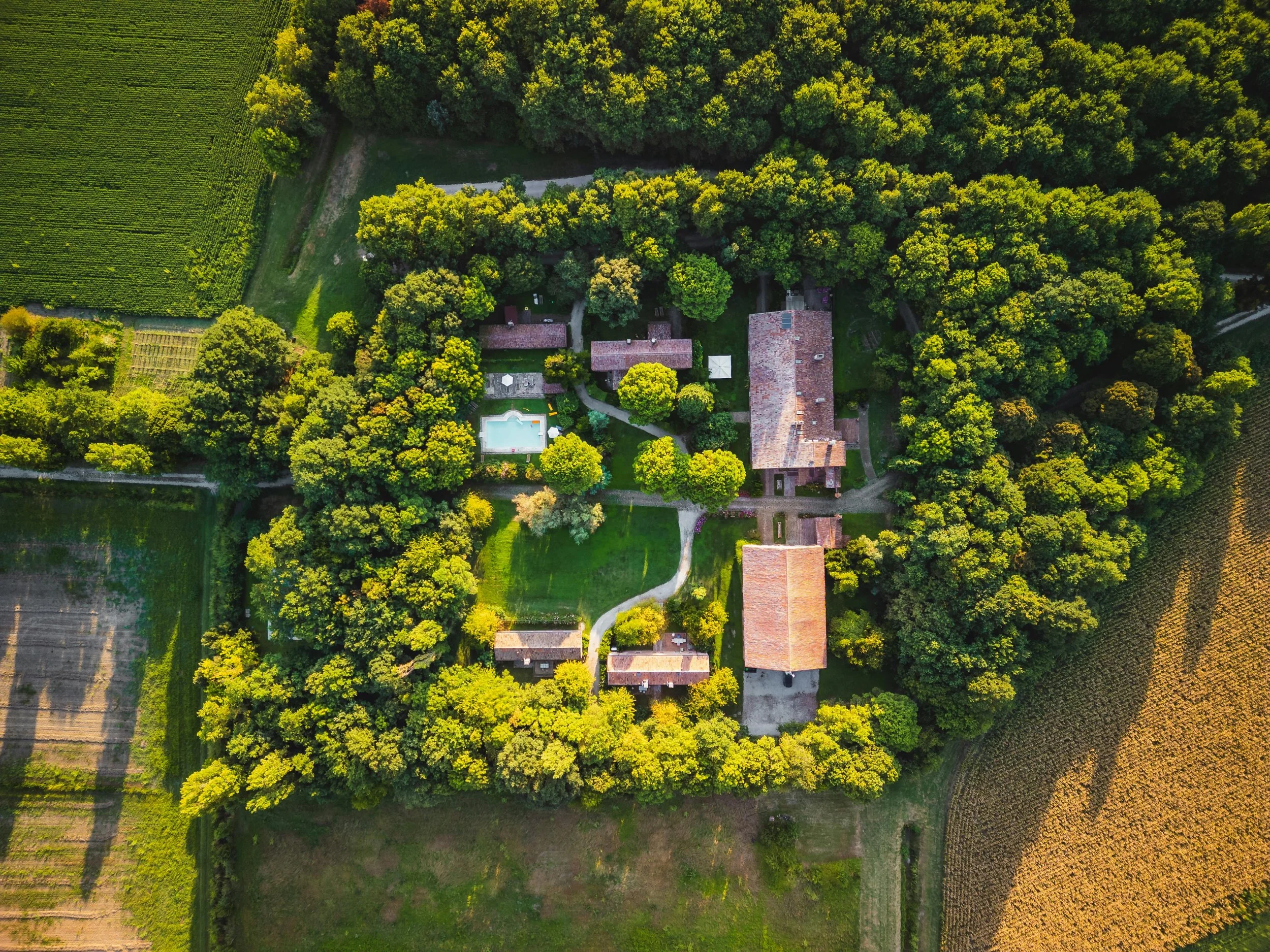 Aerial view of a large estate surrounded by trees and fields, featuring several buildings, a swimming pool, and winding paths.